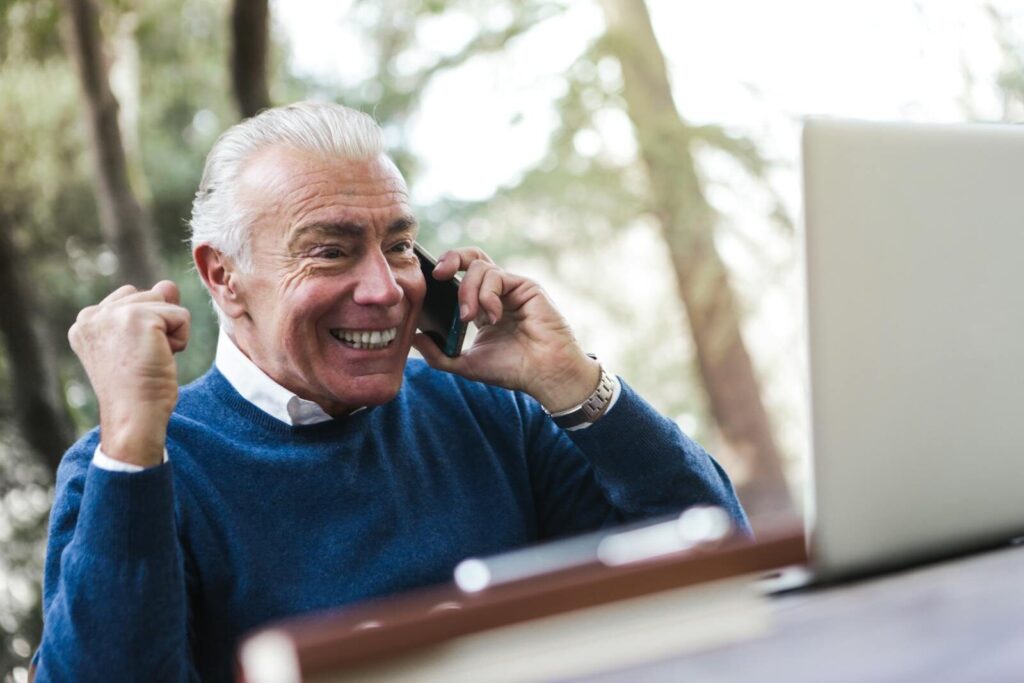 Dolfier Elderly man smiling and cheering while talking on the phone using laptop outdoors.