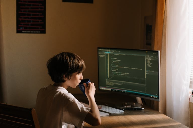 630 Area Code A young person coding at a desk with a computer and drinking from a mug.