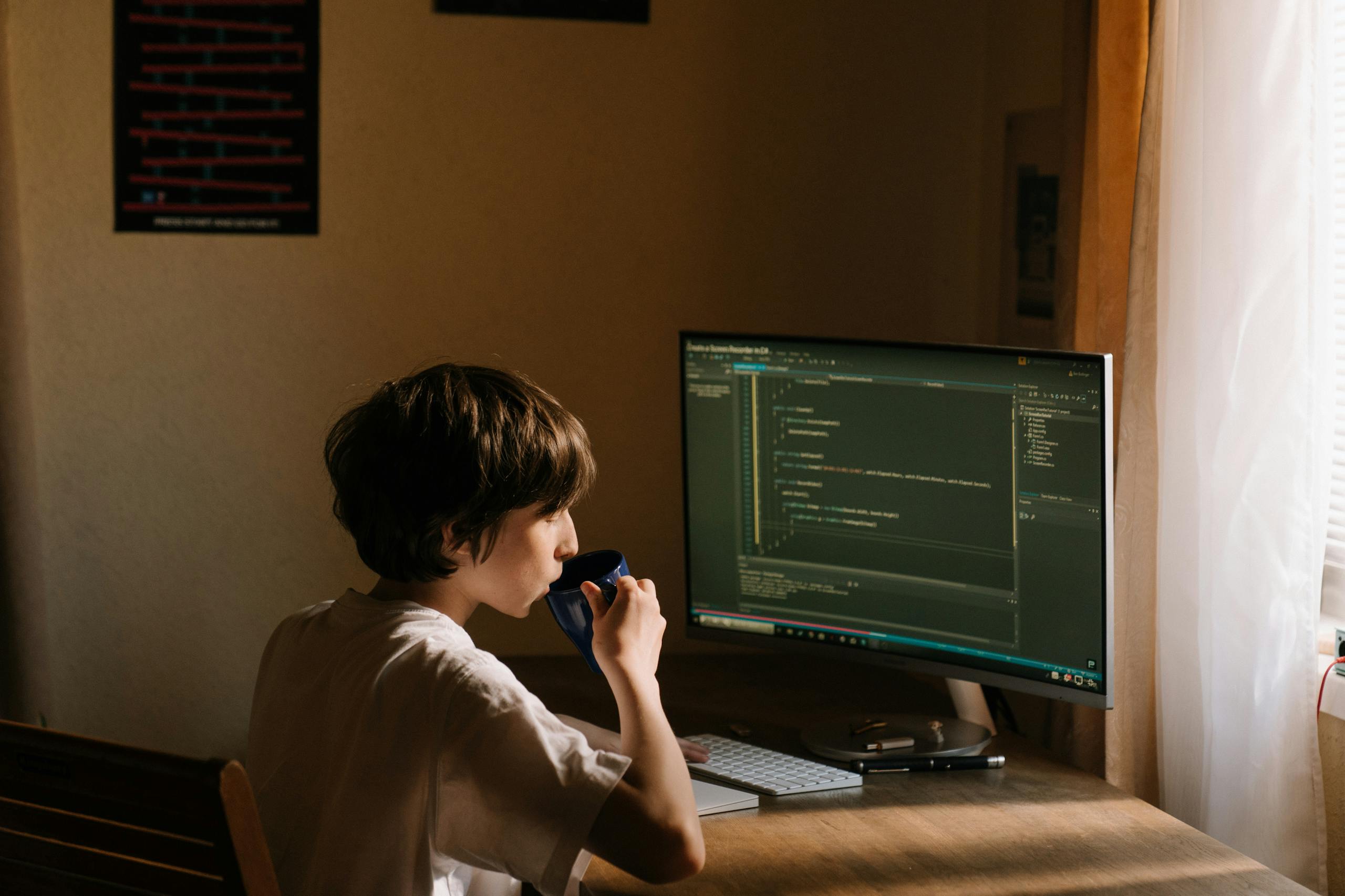 630 Area Code A young person coding at a desk with a computer and drinking from a mug.