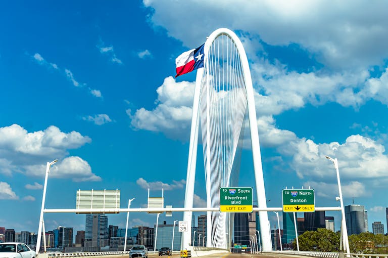Texas Hunting Forum Iconic view of Margaret Hunt Hill Bridge with Texas flag and Dallas skyline under a blue sky.