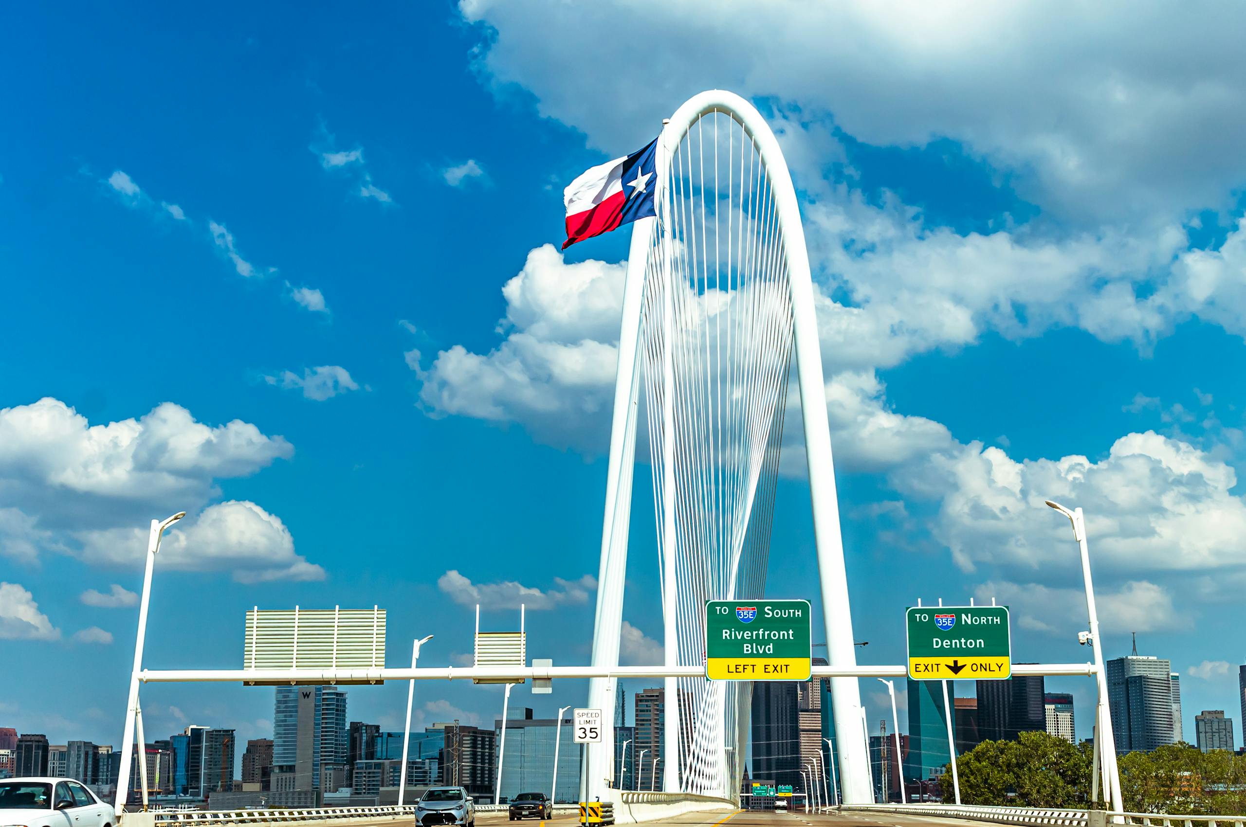 Texas Hunting Forum Iconic view of Margaret Hunt Hill Bridge with Texas flag and Dallas skyline under a blue sky.